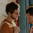 Zwei lachende Männer stehen nah beieinander auf einer Straße neben einem Blumenladen mit Blumen im Vordergrund.