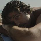 Ein Mann liegt am Strand auf einem Handtuch, sein Kopf ruht auf seinen verschränkten Armen. Er schaut direkt in die Kamera.