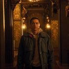 Ein junger Mann in einer blauen Jacke steht in einem opulenten, goldverzierten Gang mit warmem Licht und dekorativen Details.