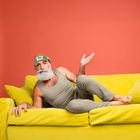 Ein Mann mit Schlafmaske liegt auf einem gelben Sofa, vor einer korallenroten Wand.