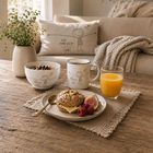 Kissen, Tasse und Schüssel beschriftet mit "ICH HAB DICH soooo LIEB!". Frühstücksszene mit Brötchen, Obst und Orangensaft.