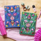 Text: "BE YOU TIFUL". Zwei mit Blumen bestickte Hüllen, eine blau, eine grün, umgeben von Schreibzubehör und Stoffen.