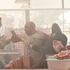 Zwei Personen kämpfen in einem verrauchten Diner. Einer liegt lachend auf dem Tresen. Essen und Getränke sind überall verstreut.