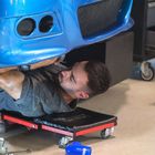 Ein Mann repariert einen blauen Autostoßfänger, liegend auf einem Rollbrett. Werkzeuge liegen um ihn herum.