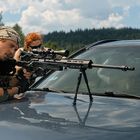Zwei Personen mit Scharfschützengewehren, die auf der Motorhaube eines Autos zielen, umgeben von bewaldeter Landschaft.