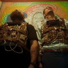 Zwei Männer mit verkabelten Westen und Timern stehen vor einem bunten Wandbild mit Gesicht. Einer trägt ein weißes Hemd.