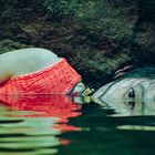 Eine Frau in rotem Oberteil liegt halb im Wasser, Kopf seitlich, ihr Auge schaut knapp über der Wasseroberfläche hervor.