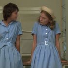 Zwei junge Frauen in hellblauen Kleidern sitzen und schauen sich an. Eine trägt zudem einen Hut. Im Hintergrund weiße Stühle.