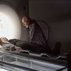 Ein alter Mann beugt sich über eine Frau, die in einer MRT-Röhre liegt. Der Raum ist schwach beleuchtet.