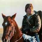 Ein Mann in Lederjacke reitet auf einem braunen Pferd mit weißer Stirn, trägt Rucksack und Patronengurte.