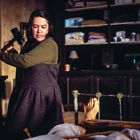 Eine Frau hält einen Hammer in einem Raum mit einem Bett. Auf dem Bett liegen Füße, die am Rahmen befestigt sind.