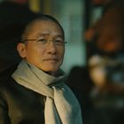 Ein älterer Mann mit Brille und grauem Schal schaut freundlich. Der Hintergrund ist unscharf mit warmen Farben.