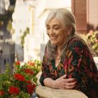 Eine ältere Frau mit weißen Haaren lächelt und lehnt an einem Balkon mit roten Blumen in einer sonnigen, mediterranen Stadt.