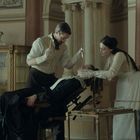 Ein Mann und eine Frau in historischer Kleidung behandeln einen Patienten in einem schicken Raum mit Säulen und Glasvitrine.