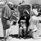 Ein Mann und eine Frau interagieren mit einem sitzenden Mann in einem Strandkorb. Gebäude im Hintergrund.