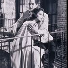 Ein Mann umarmt eine Frau auf einem Balkongeländer, beide wirken nachdenklich, umgeben von Backsteingebäuden.