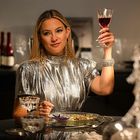 Eine Frau in silbernem Kleid hält ein Glas Rotwein hoch, sitzt an einem gedeckten Tisch.
