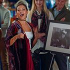 Eine Frau präsentiert ein gerahmtes Schwarz-Weiß-Foto vor einer Gruppe von Menschen. Links daneben steht eine Person und klatscht.