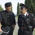Ein Priester in Schwarz mit Fahrrad spricht lächelnd mit einem uniformierten Polizisten auf einem Weg, gesäumt von Bäumen.