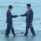 Zwei Personen stehen im flachen Wasser eines Meeres, geben sich die Hand und halten Schuhe in der anderen Hand.