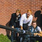 Vier Männer posieren vor einer Ziegelwand mit einer großen Waffe. Einer trägt ein blutverschmiertes Hemd.