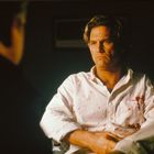 Ein Mann in weißem Hemd mit Blutflecken sitzt ernst dreinblickend in einem gedämpften Raum.