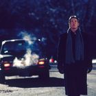 Keine Texte erkennbar. Eine Person in Mantel und Schal steht allein auf einer dunklen Straße, vor einem Auto mit dampfendem Auspuff.