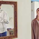 Ein Mann im Spiegel hat ein blaues Auge und knöpft ein weißes Hemd zu. Eine Frau daneben trägt ein Stirnband.