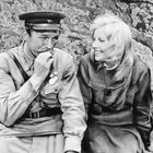 Keine sichtbaren Texte. Ein Mann in Uniform und eine Frau sitzen lächelnd vor einem Felsen.