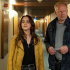 Zwei Personen in einem Flur. Eine Frau trägt eine gelbe Jacke, ein Mann hält ein Papier. Sie blicken fokussiert nach vorn.