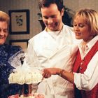 Drei lächelnde Menschen betrachten eine Hochzeitstorte mit einer Figurenverzierung oben.