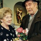 Ein Mann mit Hut lächelt, hält einen Blumenstrauß. Eine Frau mit gemustertem Kleid steht daneben, beide sind älter.