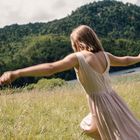 Eine Frau in einem weißen Kleid läuft durch eine blühende Wiese vor einem bewaldeten Hügel und einem See.