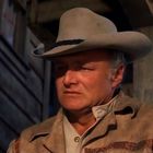 Ein Mann mit Cowboyhut und Lederjacke steht vor einem Gebäude mit Holzwand, schaut ernst, blauer Himmel im Hintergrund.