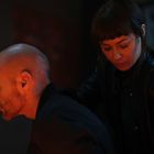 Es sind keine Texte zu sehen. Eine Frau mit Lederjacke beugt sich über einen glatzköpfigen Mann. Dramatische Beleuchtung.