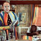 Clown mit weiß geschminktem Gesicht und buntem Anzug trocknet Geschirr, dahinter ein Affe an einem bunt gedeckten Tisch.