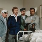 Drei Männer und ein Patient in einem Krankenhauszimmer, ein Bein hochgelegt. Gemälde an der Wand. Alle wirken ernst.