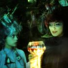 Zwei Frauen mit ungewöhnlichen Frisuren, eine hält ein leuchtendes Glas mit rotem Getränk, beide in grünem Licht.