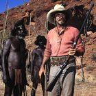 Ich kann den Text nicht lesen. Vier Männer mit Speeren und eine Person mit Cowboy-Hut und Gewehr in Wüstenlandschaft.