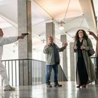 Ein Mann in Weiß zielt mit einer Pistole auf drei Personen in einem Treppenhaus, die überrascht und besorgt wirken.