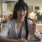 Ich kann den Text nicht lesen. Eine Frau in der Küche, lächelnd, macht ein Handzeichen und trägt ein weißes Hemd.