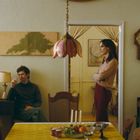 An einer Wanduhr ist es 2 Uhr. Zwei Personen, eine sitzend mit Zigarette, eine stehend mit verschränkten Armen, in einem Zimmer.