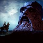 Ein Ritter auf einem Pferd betrachtet eine riesige, steinerne Gesichtsskulptur im Mondlicht.