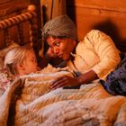 Eine Frau liegt mit einem Kind in einem hölzernen Bett, umgeben von warmem Licht. Die Stimmung ist gemütlich und fürsorglich.