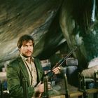 Ein Mann mit Bart in grüner Jacke hält ein Gewehr in einer Höhle, umgeben von Kisten.