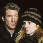 In einer nächtlichen Szene steht ein Mann mit Schal vor einem Auto, aus dessen Motorhaube Rauch aufsteigt.