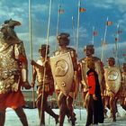 Ein Zug antiker Krieger in Rüstungen mit Schilden und Speeren am Strand, marschiert neben einer Person in rotem Gewand.