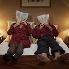 Zwei Personen sitzen auf einem Bett, tragen Plastiktüten über dem Kopf, umgeben von Nachttischen und Lampen.