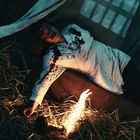 Ein Mann mit blutbefleckter Kleidung liegt auf Heu neben einem kleinen Feuer.
