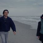 Zwei Personen gehen am Strand entlang, rechts Wellen, links Dünen, bewölkter Himmel.